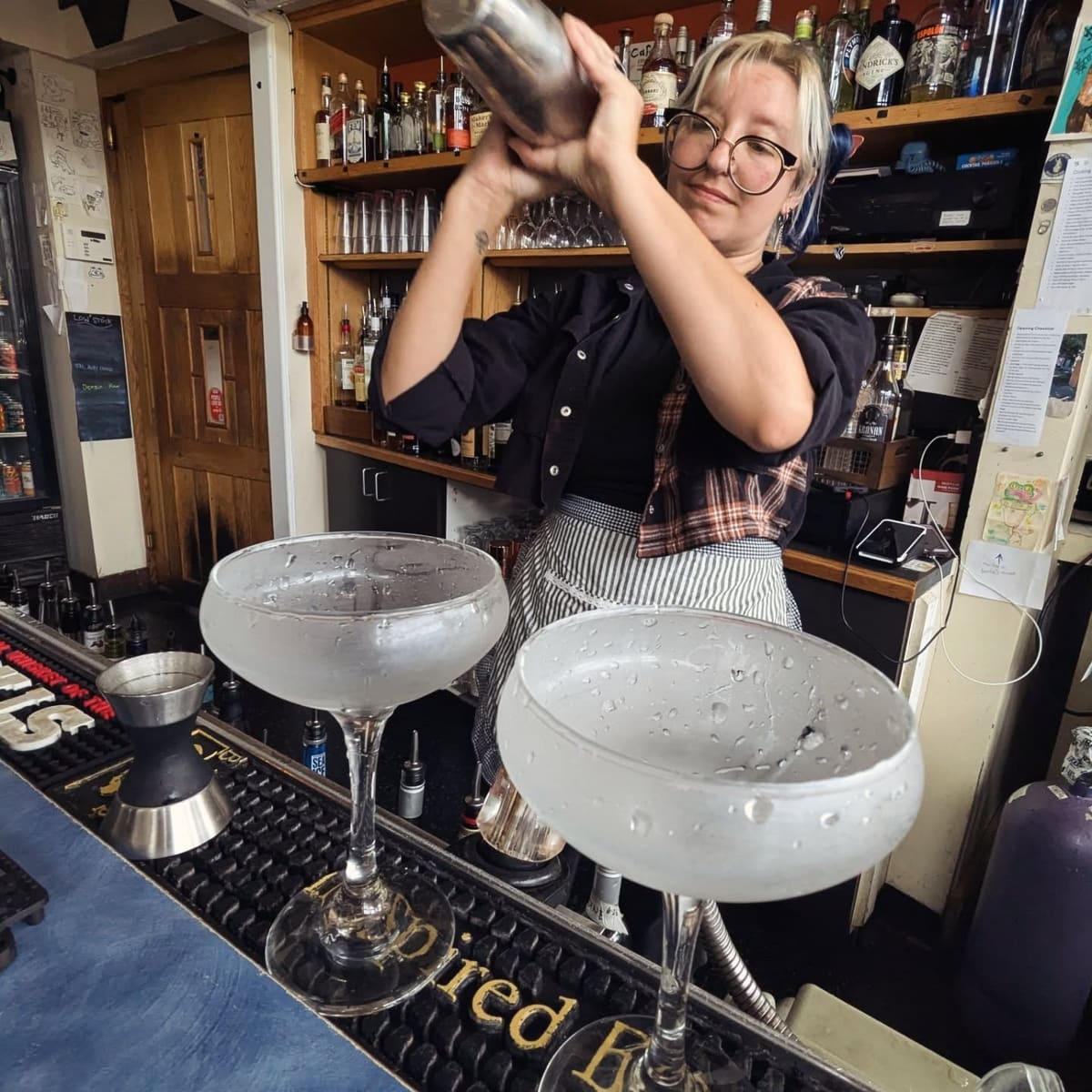 A bartender shaking a cocktail behind the bar, two coupe glasses waiting on the rail