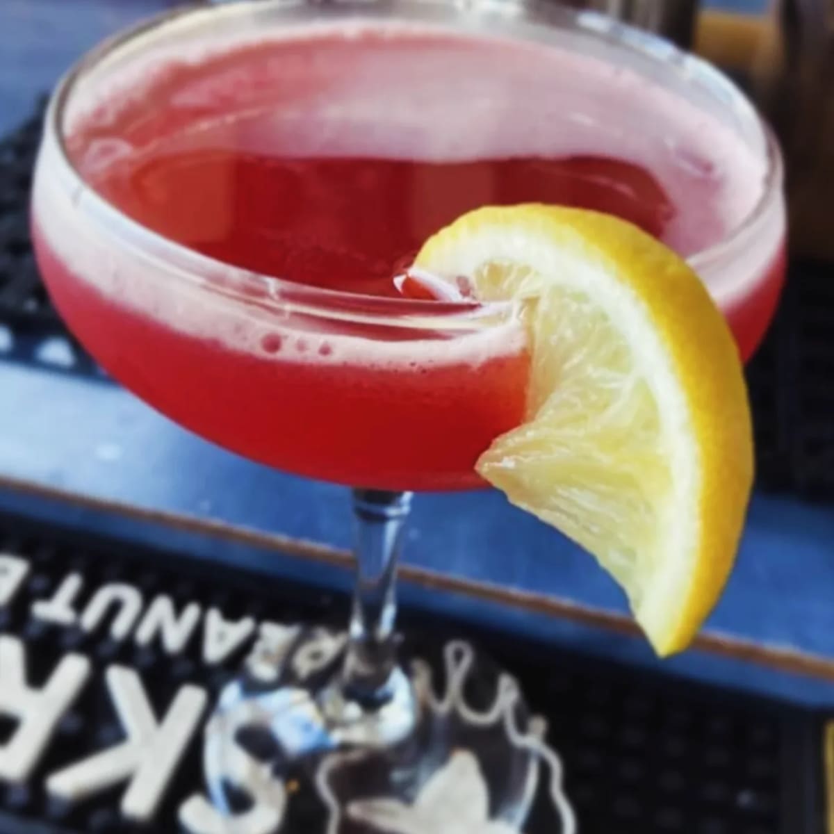 A bright red cocktail in a coupe with a lemon wheel, at the bar